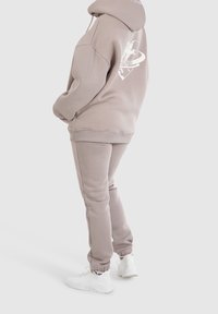 Felpa grigia chiara con un logo grafico sul retro, abbinata a pantaloni della tuta coordinati e sneakers bianche. Tessuto morbido, vestibilità rilassata.