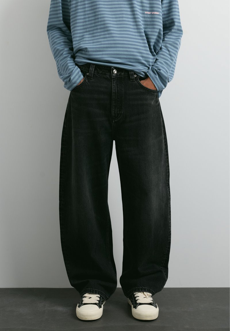 Personne portant un jean noir ample, des baskets noires et blanches, et une chemise à manches longues rayée bleue, les mains dans les poches.