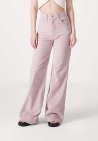 Femme portant un pantalon rose pâle taille haute à jambes larges, associé à un top court blanc et des sandales plateformes noires sur fond blanc.