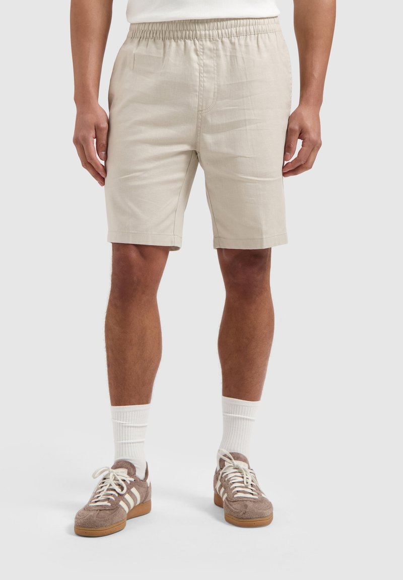 Beige shorts met een elastische tailleband, twee zijzakken, een soepele textuur en een knielange snit. Gecombineerd met bruin sneakers en witte sokken.
