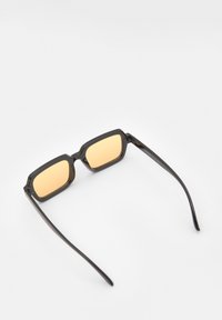 Lunettes de soleil noires rectangulaires avec des verres teintés orange et des montures en plastique élégantes. Fines branches noires droites avec une finition légèrement brillante.