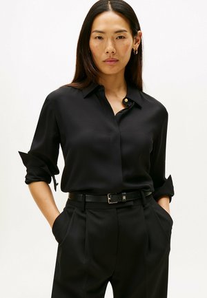 Camicia nera a maniche lunghe realizzata in tessuto morbido con colletto abbottonato e polsini arrotolati; abbinata a pantaloni neri su misura e una cintura.