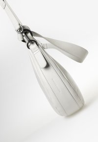 Borsa a spalla in pelle di colore grigio chiaro, con un design affusolato, tracolla regolabile e un logo intagliato discreto sul lato. Texture liscia.