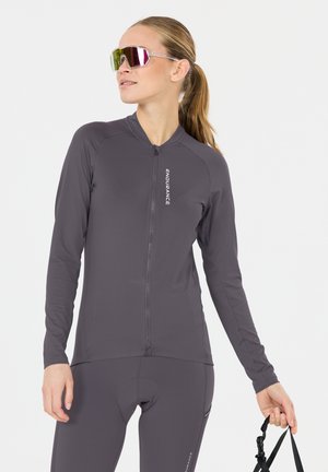 Femme avec queue de cheval portant une veste et un pantalon de cyclisme gris foncé, tenant la sangle d'un casque noir, portant des lunettes de sport réfléchissantes, regardant sur le côté.