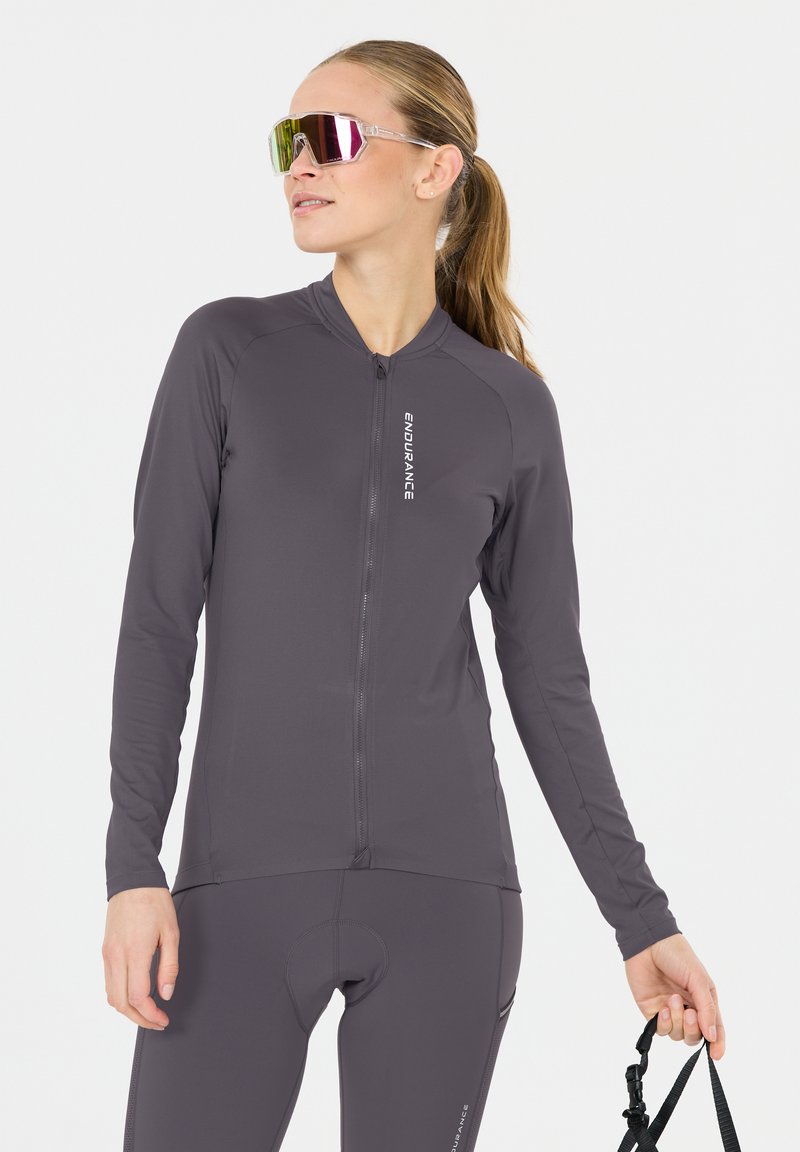 Femme avec queue de cheval portant une veste et un pantalon de cyclisme gris foncé, tenant la sangle d'un casque noir, portant des lunettes de sport réfléchissantes, regardant sur le côté.