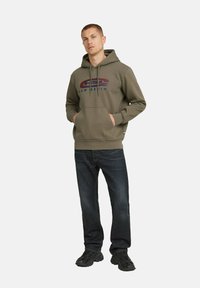 Sudadera color verde oliva con un logotipo gráfico en rojo y azul, que presenta un bolsillo frontal tipo canguro, cordones y un corte casual. Combinada con jeans oscuros.