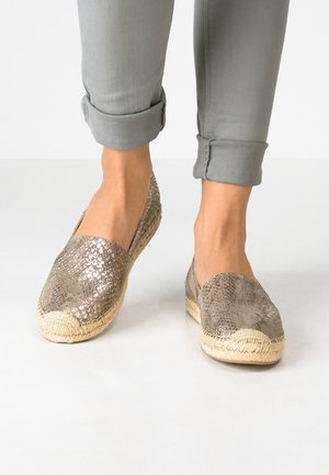 Fødder iført glitrende bronze slip-on sko med flettede såler parret med lysegrå opkørte bukser på en hvid gulv.