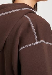 Brown hoodie with a textured fabric, featuring contrasting white stitching along the seams and hood edge. Close-up view from the side.