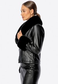 Černá kožená bunda s velkým kožíškovým límcem a kožíškově lemovanými manžetami, s přiléhavým střihem a zapínáním na zip, viděná z boku.