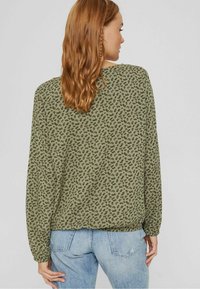 Olijfgroene blouse met een bloemenpatroon, lange mouwen en een ronde onderkant, gecombineerd met lichtblauwe jeans. Zacht materiaal, relaxte pasvorm.