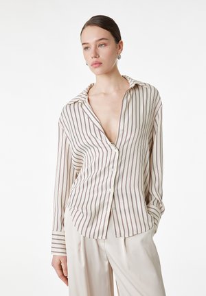 Kvinde iført cremefarvet skjorte med tynde lodrette brune striber og cremefarvede bukser med brede ben, stående med hænderne i lommerne mod hvid baggrund.