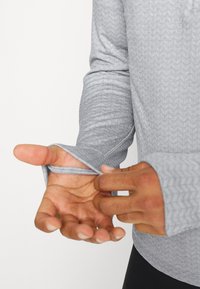 Mãos ajustando o buraco para o polegar de uma camisa de manga longa texturizada cinza-claro contra um fundo branco.