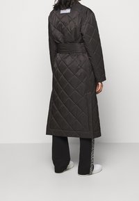 Osoba stojící zády, oblečená v dlouhém černém prošívaném kabátu s páskem, černých kalhotách s bočním textem a bílých teniskách.