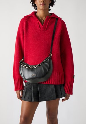 Borsa a tracolla in pelle nera con forma curva, tracolla a catena e accessori in metallo. Abbinata a un maglione rosso a maglia e a una gonna a quadri.