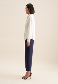 Blusa bianca a righe con colletto alto, abbinata a pantaloni eleganti blu navy e sandali con tacco neri. Design semplice e strutturato.