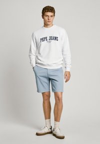 Sudadera blanca con "PEPE JEANS" impreso en azul marino, combinada con pantalones cortos azul claro. El modelo lleva zapatillas blancas con suelas marrones y calcetines claros.