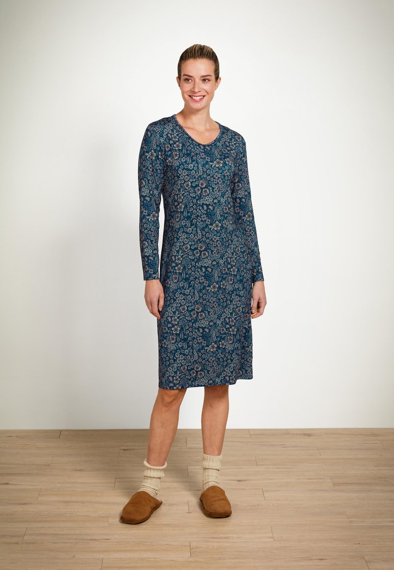 Blumenmuster, langärmliges Kleid in Dunkeltürkis mit V-Ausschnitt. Weicher, leichter Stoff, knielang, kombiniert mit braunen Schlappen.