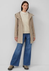 Manteau en laine beige avec capuche, fermetures éclair et poches latérales, associé à un pull en tricot crème et un jean bleu à jambes larges. Chaussures de style bottes.
