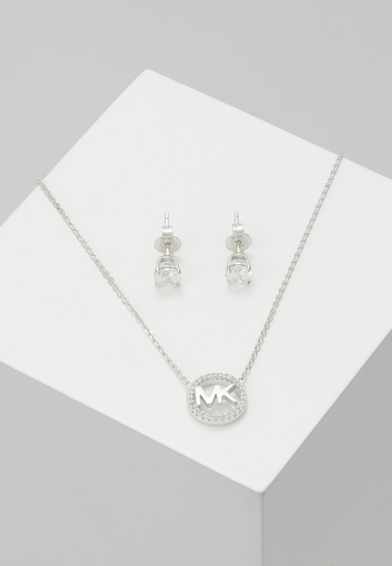 Silbernes Schmuckset mit einer Halskette, die einen kreisförmigen MK-Logo-Anhänger umgeben von klaren Steinen zeigt, sowie passenden Ohrsteckern mit klaren Steinen.