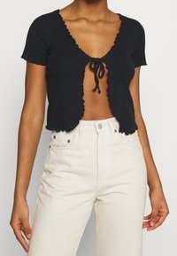 Crop top noir côtelé à manches courtes, bords festonnés et fermeture à nouer au niveau de la poitrine, associé à un pantalon taille haute beige clair.