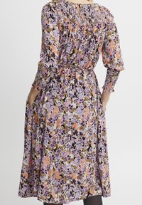 Bloemenjurk met een smock-borsten, lange mouwen en zakken. De stof toont paarse, oranje en bruine patronen op een licht achtergrond.