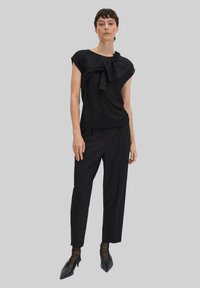 Top nero a maniche corte con dettaglio drappeggiato sul davanti, abbinato a pantaloni neri su misura e scarpe nere con punta, su uno sfondo grigio.