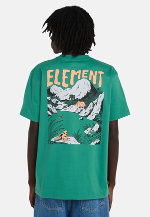Persona con capelli scuri indossa una maglietta verde con montagne, tenda, rana e piante, con la parola "ELEMENT" in grandi lettere arancioni.