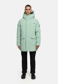 Miętowa parka z wysokim kołnierzem i regulowanym kapturem, z dwoma przednimi kieszeniami i czarnym logo na rękawie, noszona na czarnych spodniach.