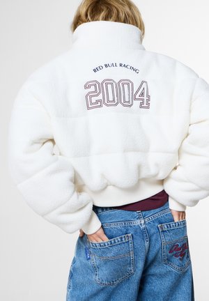 Person trägt eine weiße Fleecejacke mit "Red Bull Racing 2004" auf dem Rücken und blaue Jeans mit bestickten Gesäßtaschen.