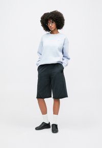 Világoskék pulóver kerek nyakkivágással, sötétszürke, térd felett érő nadrágshorts-okkal, oldalsó varrással. Fekete cipő és fehér zokni.