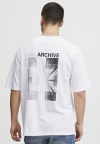 Hvid bomulds T-shirt med et grafisk print af bygninger på ryggen, med sort tekst der står "ARCHIVE." Kortærmet og løs pasform.