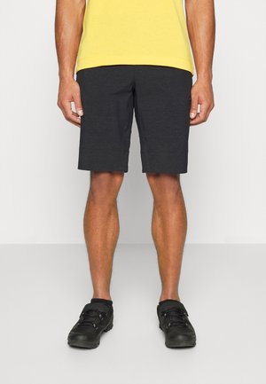 Personne portant un short noir à hauteur des genoux, des chaussures de sport noires et un t-shirt jaune, se tenant devant un fond uni clair.