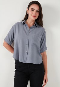 Camicetta grigia a maniche corte con bottoni e colletto, con una tasca sul petto, realizzata in tessuto leggero. Indossata con pantaloni neri.