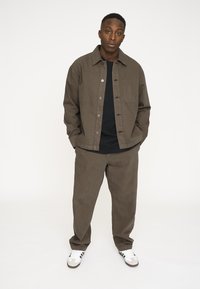 Brun bomulds skjorte med knapper og en brystlomme, parret med matchende brune bukser. Hvide sneakers med sorte striber. Minimalistisk design.