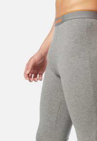 Mann in eng anliegenden grauen Leggings mit einem orangen und grauen Bund, der vor einem schlichten weißen Hintergrund steht.