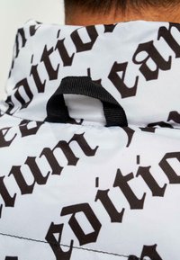 Țesătură albă cu un model de text negru mare care spune "ediție", având o buclă neagră la gât pentru suspendare. Textură netedă și ușoară.
