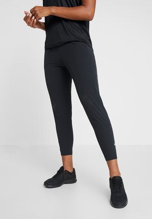 Personne portant des leggings de sport noirs et des baskets noires, debout sur un fond clair, ajustant ses mains.