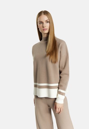 Donna con capelli lunghi e lisci che indossa un maglione beige con strisce bianche e pantaloni beige abbinati, in piedi davanti a uno sfondo bianco.