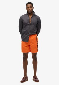Chemise grise à col, associée à un short de bain orange vif avec le logo "SUPERDRY" en noir, et des chaussures à lacets marron.