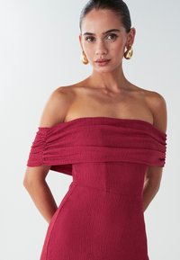Rotes, schulterfreies Kleid aus strukturiertem Stoff, mit gerafften Ärmeln und einer taillierten Silhouette. Es werden goldene Ohrhänger getragen.