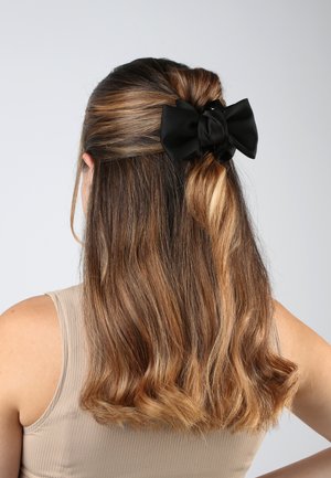 Fermaglio in satin nero attaccato a una coda di cavallo, in contrasto con i capelli ricci castani, su uno sfondo chiaro e semplice.