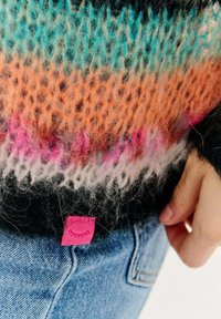 Sweter z dzianiny w kolorowe paski w odcieniach turkusu, pomarańczy, różu i czerni; posiada różową metkę na dolnym lewym boku.