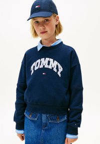 Sudadera azul marino con la palabra "TOMMY" impresa en blanco, combinada con una camisa de cuello azul claro y jeans de mezclilla con dos bolsillos traseros.
