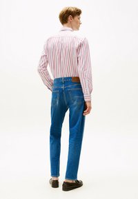 Vista trasera de una persona que lleva una camisa a rayas rosa y blanca y jeans azules con un parche de cuero, complementados por mocasines oscuros.