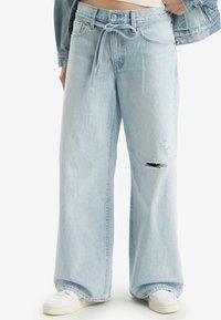 Jeans in denim azzurro chiaro a gamba larga con vita alta, dotati di cintura con nodo e dettaglio strappato sulle ginocchia. Abbinati a sneakers bianche.