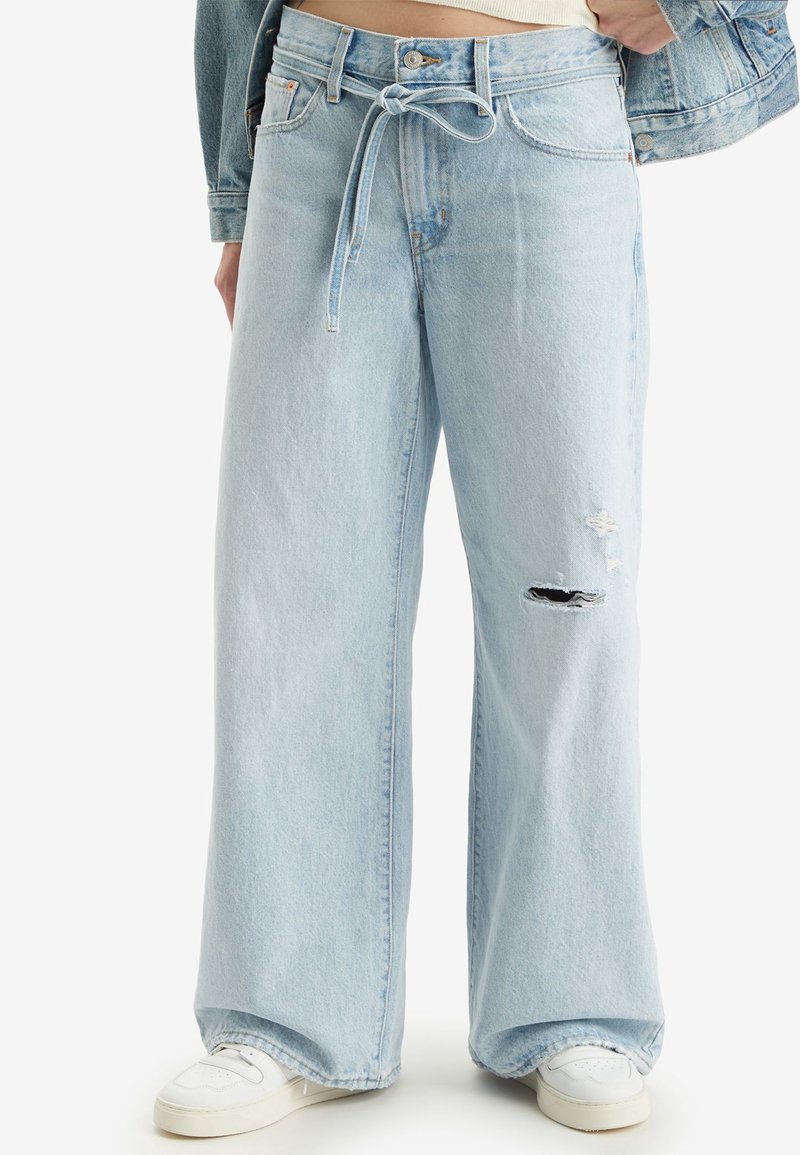 Jeans in denim azzurro chiaro a gamba larga con vita alta, dotati di cintura con nodo e dettaglio strappato sulle ginocchia. Abbinati a sneakers bianche.