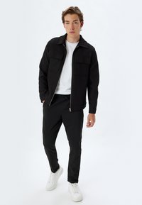 Sort ribbet jakke med to forlommer, hvid t-shirt, sorte bukser og hvide sneakers, stående foran en ensfarvet baggrund.