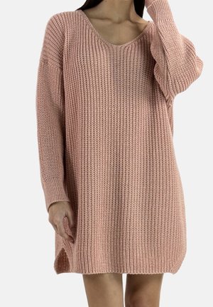 Femme portant une robe-pull en maille oversize rose poudré avec longues manches et fentes latérales, debout devant un fond uni.