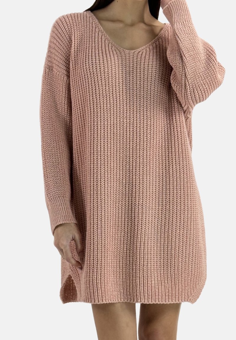 Femme portant une robe-pull en maille oversize rose poudré avec longues manches et fentes latérales, debout devant un fond uni.