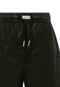 Pantalon synthétique noir et brillant avec une taille élastique, des accents de cordon de serrage et une étiquette Calvin Klein au centre.